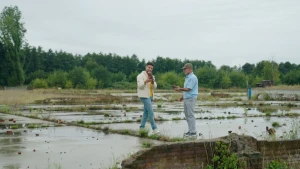 Een foto van twee mensen die buiten staan te praten. Ze staan op betonnen platen met onkruid ertussen. Ze hebben stenen in hun handen.