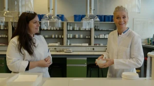 Een foto van presentator Janouk en een andere persoon, die achter een tafel staan. Ze hebben witte werkkleding aan en staan in een ruimte met heel veel potjes en doosjes. Janouk heeft een kommetje vast.