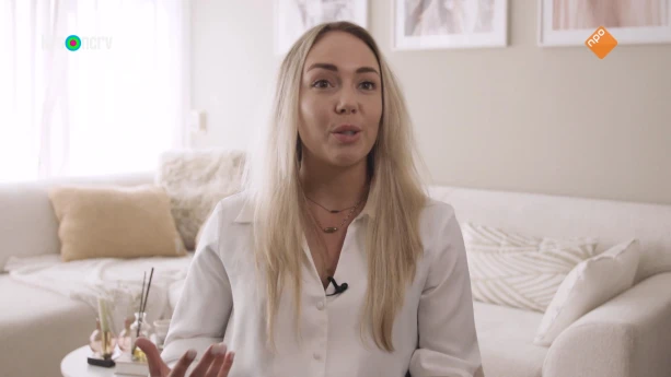 Een foto van een vrouw die in een woonkamer zit. Ze praat tegen iemand die naast de camera zit. Achter haar s een grote bank te zien, een groot raam en schilderijen aan de muur.