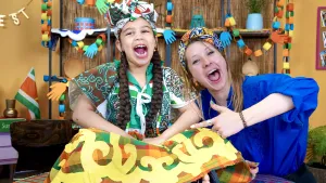 Een foto van Rosalie en een meisje die met open mond een uitroep doen, beiden met een veelkleurige doek om hun hoofd. Het meisje zit op een tafel. De omslagdoek om haar rok heeft gekleurde oranje, groene en rode vierkantjes en gekrulde gele lijnen. 