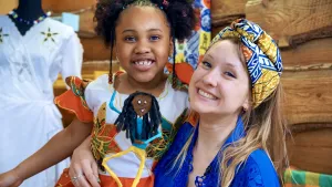 Een foto van Rosalie en een meisje. Rosalie heeft haar arm om het meisje heen en heeft in haar andere hand een dansend poppetje met een blij gezicht, lang, zwart haar, lange benen en met één arm in de lucht. Het draagt een gele broek en een blauwe trui.