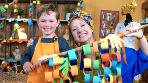 Een foto van Rosalie en Sem die een tak vasthouden, waar papierstrookjes overheen hangen met oranje, gele, groene en blauwe kleuren en af en toe een papieren hand ertussen. Om ieder stookje hangt een ander strookje heen en zo is het een lange slinger.