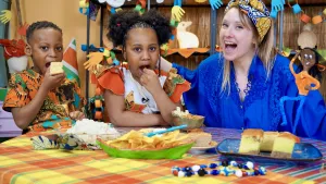Een foto van Rosalie met een blauwe blouse en een doek om haar hoofd en een jongen en een meisje in rood en oranje gekleurde kleding. Ze zitten achter een tafel met vier bakjes en een bordje vol met hapjes. De kinderen steken eten in hun mond op de foto. 