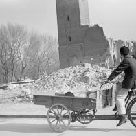 Zwartwit foto van een persoon op een bakfiets die langs een gebombardeerd gebouw rijdt.