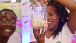 Een foto van de twee presentatoren Tirsa en Eva. Tirsa kijkt enthousiast de camera in. Eva kijkt blij naar de glazen pot in haar hand. In de pot zit rijst met gekleurde bolletjes. 