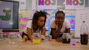 Een foto van 2 mensen die op een tafel leunen. Op de tafel staan blikjes frisdrank, en glazen potten met water en frisdrank. De mensen kijken naar de pot met cola.