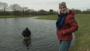 Een foto van de presentator die naast een meer staat en een duiker die in het meer zit. De duiker heeft een speciaal pak aan om onder water te kunnen zwemmen en ademen..