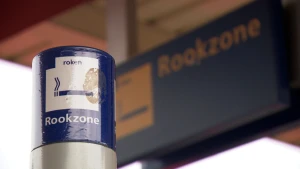 Een foto van een stalen paal waar aan de zijkant de woorden "roken" en "Rookzone" te lezen zijn met het plaatje van een sigaret erop. Op de achtergrond is ook een groot blauw bord zichtbaar met dezelfde teks en een plaatje van een sigaret erop.