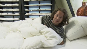 Een foto van presentator Pascal die onder een wit dekbed in een bed ligt. Ook ligt er een wit kussen zonder hoes. Op de achtergrond staan kasten met planken waar andere witte dekbedden opliggen.