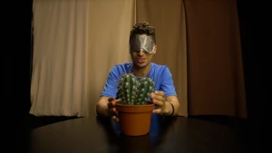 Een foto van presentator Nizar die achter een tafel zit. Nizar heeft een oogmasker op en voelt met zijn handen aan de stekels van een cactus die op de tafel staat. Achter Nizar hangt een lichtbruin gordijn.
