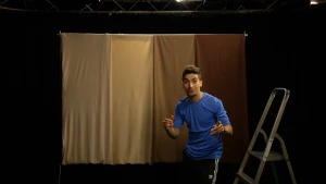 Een foto van presentator Nizar in een donkere ruimte. Naast Nizar staat een trap op de grond. Achter Nizar hangt een lichtbruine gordijn aan een stang.