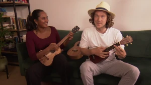 Een foto van presentator Pascal en zangeres en muzikant Indra van de Bilt die met een ukelele in de hand op de bank zitten en op de ukelele spelen. Een ukelele is een soort kleine gitaar. Pascal heeft een lichtgele hoed op met een zwarte band erom.