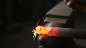 Een foto van een stalen blok waarop een verhitte staaf ijzer met een roodgloeiende bovenkant ligt. De gloeiende bovenkant wordt in vorm geslagen met een hamer. De punt van de hamer is nog net zichtbaar in de rechterbovenhoek van de foto. 
