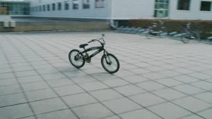 Een foto van een zwarte kinderfiets van het type BMX die op een groot plein staat. Op de achtergrond zijn een deel van een gebouw en een fietsenrek met een paar fietsen erin te zien.