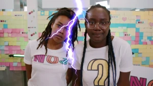 Een foto van presentatoren Eva en Tirsa die een wit t-shirt dragen. Op dat van Eva staat "NOJ" en op dat van Tirsa een vraagteken. Voor hen in het midden is een wit-paarse bliksemschicht zichtbaar. Op de achtergrond hangen gekleurde briefjes.