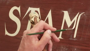 Een foto van een hand die op een bruine houten plank witte letters met een dunne kwast schildert en ook een stok met een lapje stof vastheeft om de plank niet te raken. Letter "M" wordt op de foto geschilderd, samen met letters ervoor is "Stam" te lezen.
