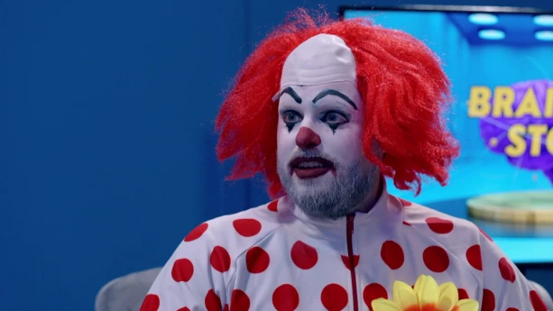 Een foto van een clown met een rode pruik op zijn hoofd die een wit vest met rode bolletjes erop aanheeft. De clown heeft een wit beschilderd gezicht, waarop de neus en lippen rood geschilderd zijn en de wenkbrauwen en de randen om de ogen zwart. 
