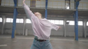 Een foto van een danseres die in een lege magazijnhal urban contemporary, een mix van verschillende dansstijlen, danst. Ze kijkt met haar schuin omhoog geheven hoofd naar haar rechts geheven rechterhand en strekt de linkerhand links naar achteren.