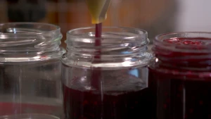 Een foto van drie glazen potjes. Het rechterpotje is geheel gevuld met Rode Jam. Het middelste potje wordt op de foto gevuld met rode jam door een spuitmond van een machine. De machine is niet te zien op de foto. Het linkerpotje is nog helemaal leeg.