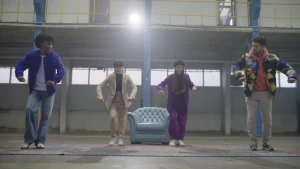 Een foto van een presentator die in een lege hal van een magazijn met drie dansers de Amerikaanse dans locking danst.  Ze staan in een houding met beide armen naar voren en de vuisten gebald en tillen hun rechterbeen een heel klein beetje op. 