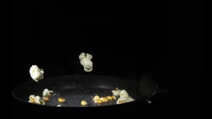 Een foto van een zwarte koekenpan met popcorn erin. Er liggen losse maïsstukjes in uit het binnenste van de popcorn en ook stukjes hele popcorn. Op de foto springen twee stukjes hele popcorn omhoog uit de pan. De achtergrond van de foto is zwart.