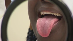 Een foto van een vrouw die haar tong uitsteekt. De mond, een klein stukje van de neus en een stuk van de staart van het haar van de vrouw zijn zichtbaar. Dit beeld is te zien vanuit een spiegel die de vrouw in haar hand heeft.