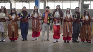 Een foto van een rij mannen en vrouwen die de Turkse volksdans Halay dansen in kleurige, traditionele Turkse kleding. De presentator van het programma staat in het midden van de rij. Ze staan dichtbij elkaar, de handen in een gebalde vuist tegen elkaar.