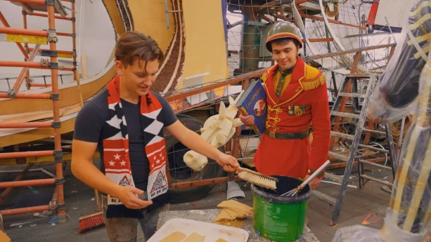 Een foto van twee mensen bij een tafeltje. Een jongen heeft gewone kleding aan en doopt een grote kwast in een emmer met lijm. De andere jongen heeft een speciaal pak aan van gekleurde legerkleding. Ze staan in een ruimte met ladders en een grote wagen.