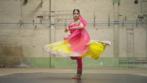 Een foto van een vrouw die de Kathak danst. Ze staat op één voet en haar rok draait rond. De vrouw draagt Indiase kleding, bestaande uit een gele rok, rode bovenkleding, oorbellen, een ketting, een armband, een enkelband en een sieraad op het voorhoofd.