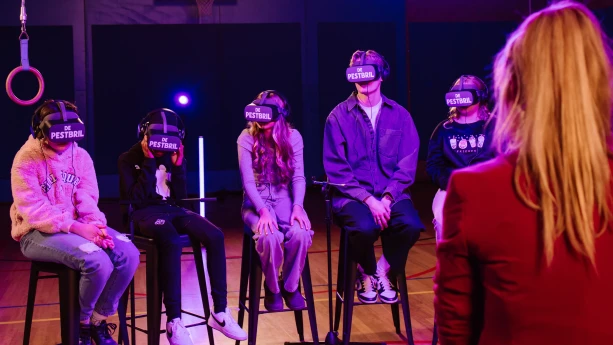 Een foto van influencer Duncan Tromp en vier kinderen die op een hoge kruk in een gymzaal zitten. Ze hebben een VR, Virtual Reality, bril op met de tekst "De Pestbril" erop geschreven. Rechts vooraan is de rug van presentator Anne-Mar te zien.