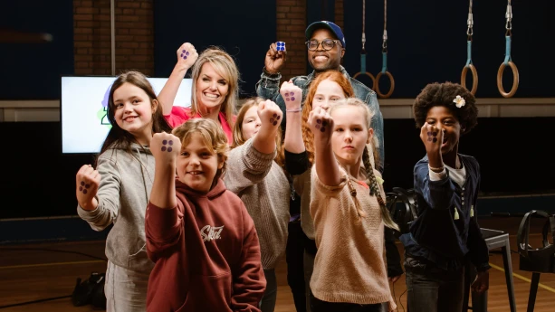 Een foto van komiek Jandino Asporaat, Anne-Mar en zes kinderen die staan in een gymzaal. Jandino houdt een vierkant wit velletje met vier blauwe puntjes omhoog. Anne-Mar en de kinderen heffen hun gebalde vuist met daarop vier zwarte stippen als vierkant.