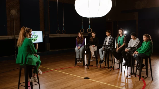 Een foto van zangeres, actrice en presentatrice Stefania Liberakakis die tussen vijf kinderen inzit in het midden van een gymzaal. De kinderen en Stefania zitten op een hoge kruk rechts en links in beeld zit presentator Anne-Mar ook op een hoge kruk.