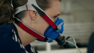 Een foto van de zijkant van het gezicht en de schouder van presentator Petra Grijzen. Ze draagt een blauw masker dat met een roodwitte band vastzit aan haar hoofd. Van de voorkant van het masker lopen twee slangetjes naar een niet-zichtbaar meetapparaat.