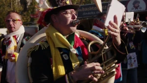 Een foto waarop muzikanten en één carnavalsvierder staan. Groot in beeld is een man in carnavalskleding die het muziekinstrument Tuba bespeeld. Hij kijkt naar bladmuziek in zijn hand tijdens het spelen. De man draagt een hoed met veren en twee sjaals. 