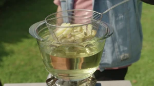 Een foto waarin iemand een klein mandje met patat in een glazen kom met olie doet. Onder de kom staat een gasbrander. De foto is genomen buiten op een grasveld. Van de persoon op de foto is geen gezicht te zien, alleen een gedeelte van de kleding.