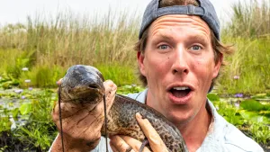 Een foto van presentator Freek die vlak voor de camera staat en een dier vasthoudt die in het water leeft.