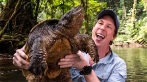 Een foto van presentator Freek die aan een oever in een tropisch gebied een grote bijtschildpad vasthoudt. 