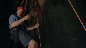 Een foto van presentator Freek die in een grot naar beneden gaat met een touw. Hij legt wat uit in de camera.