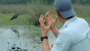 Een foto van presentator Freek die aan een oever staat met zijn rug naar de camera toe. Hij kijkt naar een Schoenbekooievaar. Dat is een hele grote vogel. 