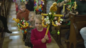 Een foto van een rij kinderen die in het gangpad tussen de kerkbanken lopen. De kinderen dragen een palmpasenstok in de vorm van een kruis. Op de stok staat een broodhaantje en aan de stok hangen naast een palmtak veel snoepjes.