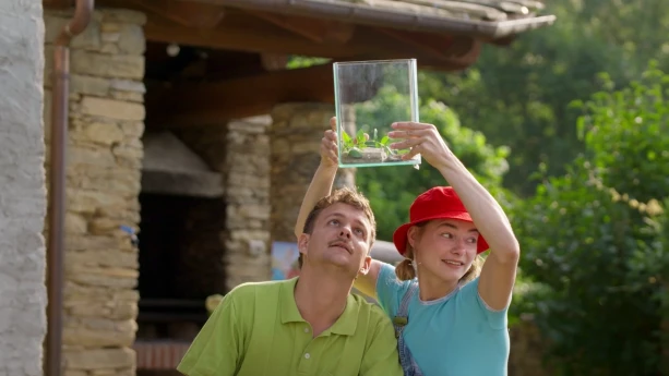 Een foto van presentatoren Jurre en Emma. Emma houdt een bak waar je doorheen kan kijken boven het hoofd van Jurre zodat hij aan de onderkant kan kijken. In de bak zit een duizendpoot samen met een plantje en een steen.