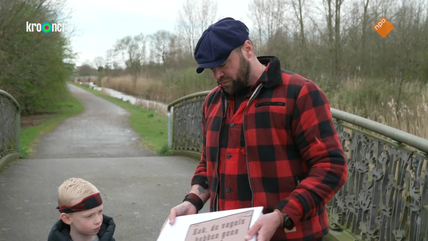 Een foto van presentator Tim die samen met een jongetje op een brug staat. Tim heeft een witte houten bord in z'n hand met een tekst erop.