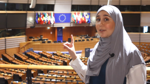 Een foto van een presentator die in de zaal van het Europees Parlement in Brussel staat. De presentator wijst omhoog naar de Europese vlag die op de achtergrond zichtbaar is.