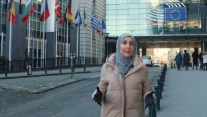 Een foto van een presentator die op straat staat met op de achtergrond een gebouw van de Europese Unie. Naast de Europese vlag die op het gebouw staat, staan aan de linkerkant ook vlaggenmasten met tien vlaggen van diverse Europese landen.