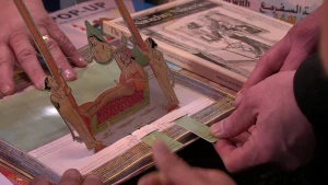 Een foto van een pop-up boek dat op een marktkraam staat. Op de foto trekt iemand met zijn hand aan het strookje van het pop-up boek om het plaatje naar boven te laten springen. Het is een getekend plaatje met een seksstandje van een man met een vrouw.