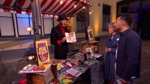 Een foto van oud-leraar Nederlands Willard Mans die buiten achter een marktkraam vol met graphic novels staat. Presentator Özcan Akyol staat samen met een mevrouw voor de kraam. 