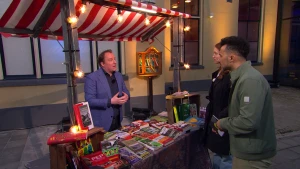 Een foto sportverslaggever Mark van Rijswijk die buiten achter een marktkraam vol met voetbalboeken staat. Presentator Özcan Akyol staat samen met een mevrouw voor de kraam.