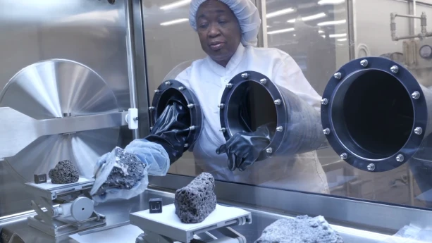 Een foto van een onderzoeker die in een luchtdichte kast een stuk maansteen plaatst die meegenomen is door een astronaut van de maan. Er zijn in de kast ook twee andere stukken maansteen zichtbaar.