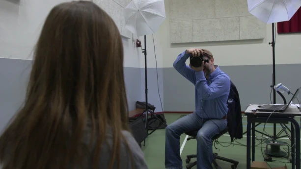 Een foto van een fotograaf die een foto neemt van een persoon die achterstevoren zichtbaar is. Naast de fotograaf staan lampen die in een fotostudio gebruikt worden en een tafel met daarop een laptop. De foto is genomen in een gymlokaal.