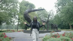 Een foto van presentator Tirsa die wat uitlegt voor de camera over het slavernijmonument op de achtergrond.